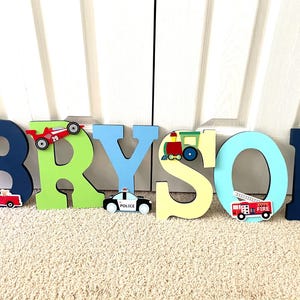 Puede incluir: Letras de madera de colores que deletrean "BRYSON", decoradas con motivos de veh&iacute;culos. Incluye un coche de carreras rojo, un cami&oacute;n rojo, un coche de polic&iacute;a, un tren y un cami&oacute;n de bomberos.