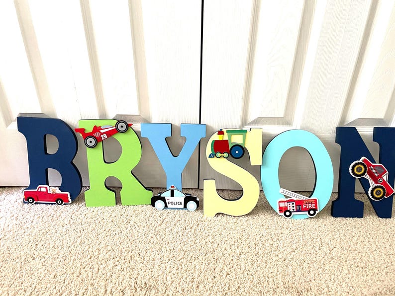 Puede incluir: Letras de madera de colores que deletrean "BRYSON", decoradas con motivos de veh&iacute;culos. Incluye un coche de carreras rojo, un cami&oacute;n rojo, un coche de polic&iacute;a, un tren y un cami&oacute;n de bomberos.