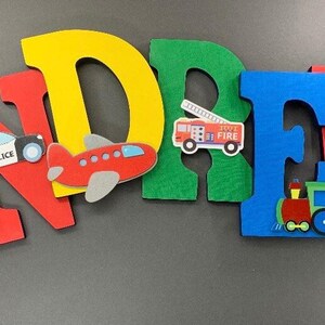 Puede incluir: Letras de fieltro de colores que deletrean un nombre, con recortes de veh&iacute;culos adjuntos. Las letras son rojas, amarillas, verdes y azules. Los veh&iacute;culos incluyen un coche de polic&iacute;a, un avi&oacute;n, un cami&oacute;n de bomberos y un tren.