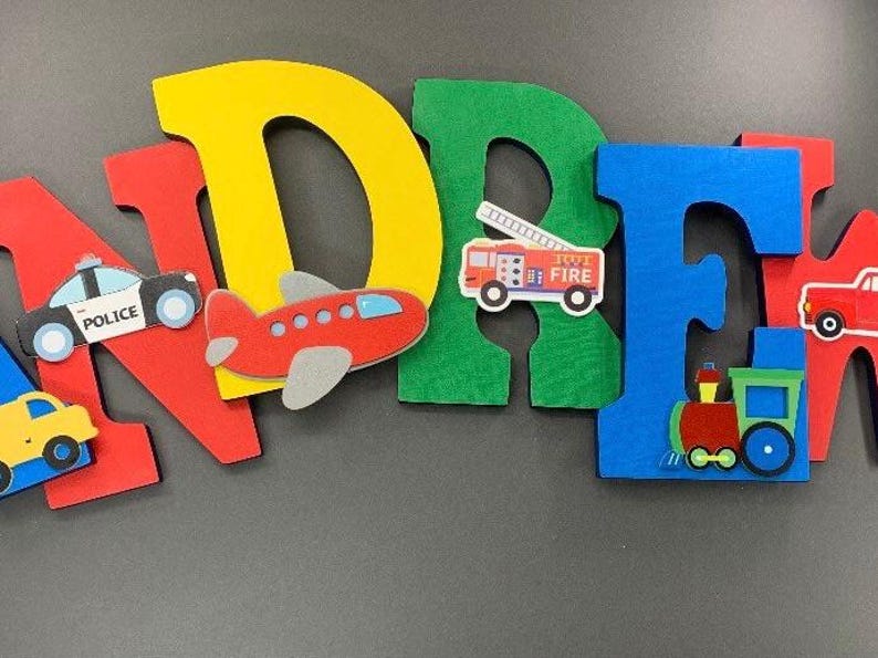 Puede incluir: Letras de fieltro de colores que deletrean un nombre, con recortes de veh&iacute;culos adjuntos. Las letras son rojas, amarillas, verdes y azules. Los veh&iacute;culos incluyen un coche de polic&iacute;a, un avi&oacute;n, un cami&oacute;n de bomberos y un tren.