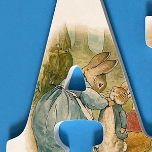 May include: A white wooden letter A with a picture of Peter Rabbit and his mother on it. The letter is on a blue background.