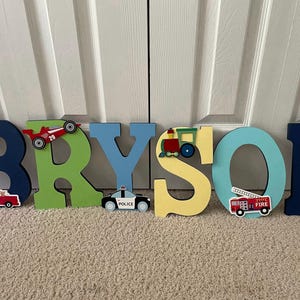 Puede incluir: Letras de madera que deletrean "BRYSON" en varios colores, cada una decorada con recortes tem&aacute;ticos de veh&iacute;culos. Incluye un cami&oacute;n rojo, un coche de carreras, un coche de polic&iacute;a, un tren, un cami&oacute;n de bomberos y otro coche de carreras.