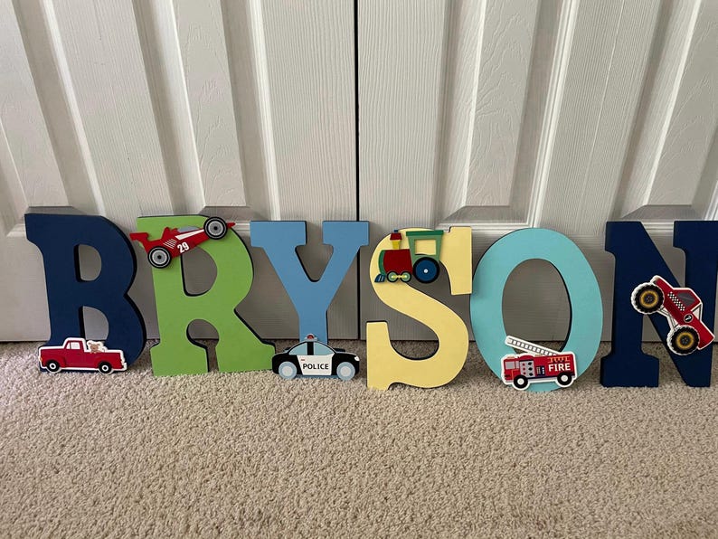 Puede incluir: Letras de madera que deletrean "BRYSON" en varios colores, cada una decorada con recortes tem&aacute;ticos de veh&iacute;culos. Incluye un cami&oacute;n rojo, un coche de carreras, un coche de polic&iacute;a, un tren, un cami&oacute;n de bomberos y otro coche de carreras.