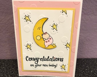 May include: A pink and yellow baby shower card with a white background. The card features a cartoon baby sleeping on a yellow crescent moon with stars. The text on the card reads "Congratulations on your new baby!"