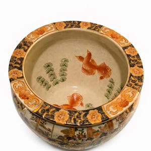 May include: A round, ceramic Japanese fish bowl with a cream-colored interior featuring two orange goldfish and green plant illustrations. The rim is decorated with orange floral patterns and black accents. The exterior has a detailed landscape design.
