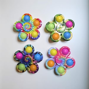 May include: Four flower-shaped fidget toys with colorful push-pop bubbles. Two have tie-dye patterns, and the others have green and yellow camouflage patterns. Each toy has five bubbles in various colors, including pink, blue, orange, and yellow.