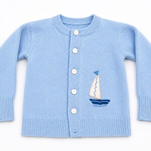 May include: A light blue long-sleeve knit cardigan featuring a crew neckline, ribbed cuffs and hem, and five white buttons down the front. An embroidered sailboat design with a white sail and dark blue hull is positioned on the lower right side.