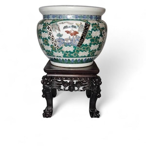 May include: A large, ornate ceramic planter with a detailed landscape design in green, white, and blue, set on a dark wooden stand. The stand features carved dragon details. The planter is a decorative piece.