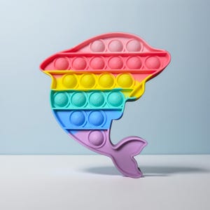 May include: A colorful dolphin-shaped pop-it toy. The toy features rows of bubble-like circles in shades of pink, red, yellow, green, blue, and purple. The toy is designed with a dolphin's head, body, and tail.