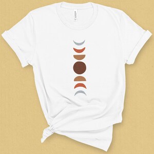May include: White t-shirt with a vertical design of a moon phases graphic in shades of gray, brown, and orange.