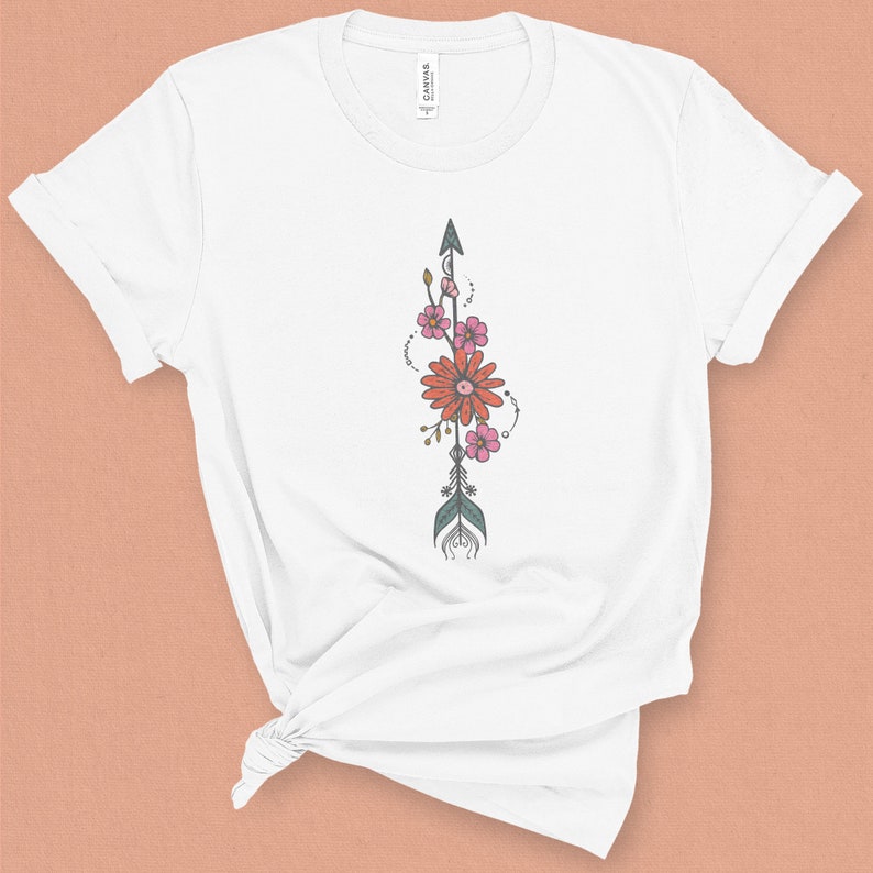 May include: White t-shirt with a floral arrow design. The arrow is green and gray with pink and orange flowers.