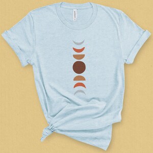 May include: A light blue t-shirt with a graphic of a stylized moon phases design in shades of gray, brown, and orange.