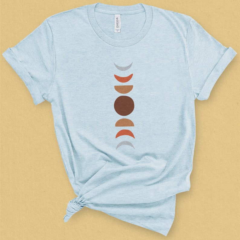 May include: A light blue t-shirt with a graphic of a stylized moon phases design in shades of gray, brown, and orange.