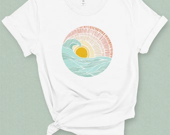 May include: White t-shirt featuring a circular graphic of a stylised wave and sun in shades of blue, yellow, pink and orange.