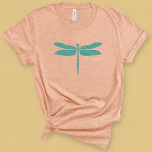 May include: A light peach colored t-shirt with a teal dragonfly graphic design. The dragonfly has a detailed design with small circles throughout the wings.