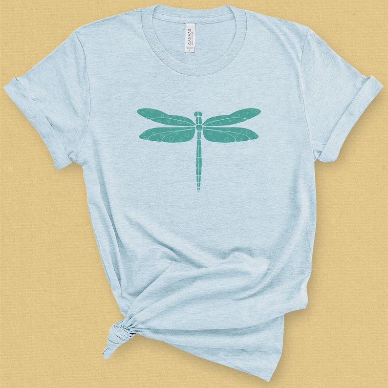 May include: A light blue t-shirt with a teal dragonfly graphic. The dragonfly has a detailed design with small circles throughout its wings.
