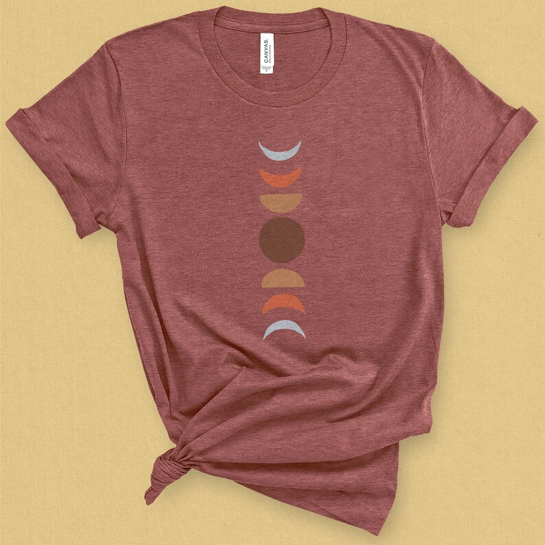 May include: A maroon t-shirt with a graphic of a stylized moon in various phases. The moon phases are represented by a light blue crescent, an orange crescent, a brown circle, a tan crescent, and a light blue crescent.