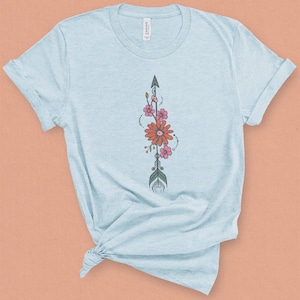 May include: A light blue t-shirt with a floral arrow design. The arrow is black and has a green feather. The flowers are pink and orange.