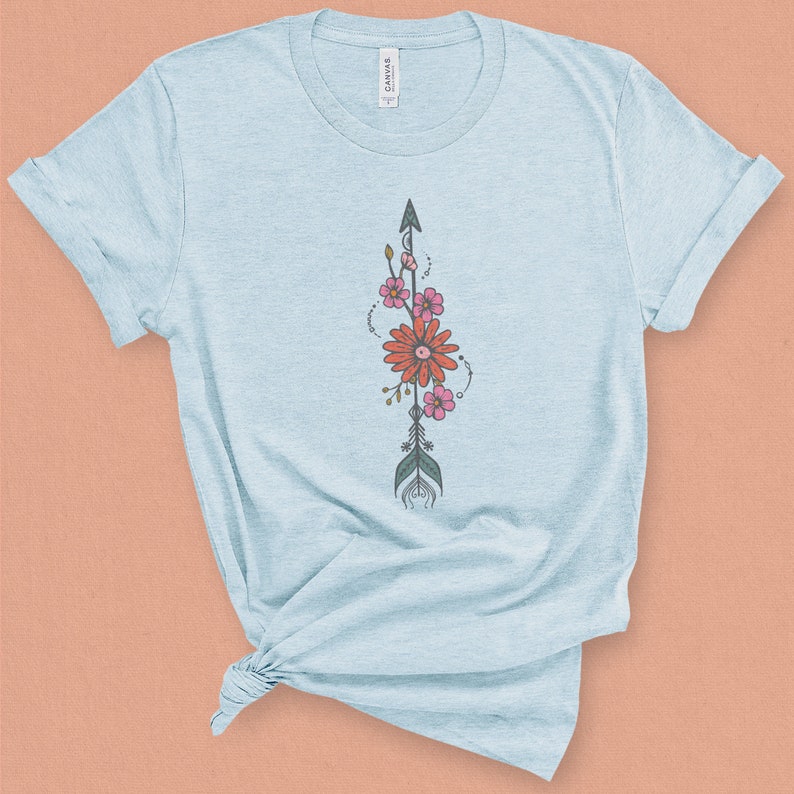 May include: A light blue t-shirt with a floral arrow design. The arrow is black and has a green feather. The flowers are pink and orange.