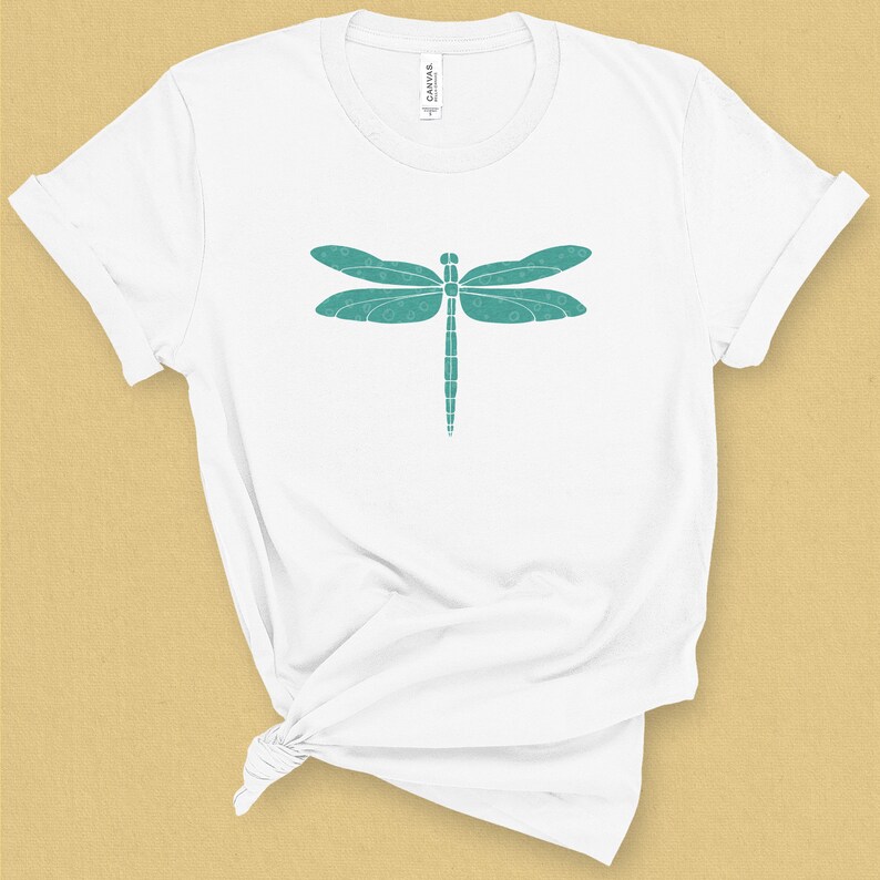 May include: White t-shirt with a teal dragonfly graphic. The dragonfly is made up of small circles.