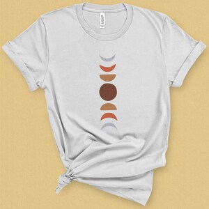 May include: White t-shirt with a graphic of a vertical line of abstract moons in shades of gray, red, orange, and brown.