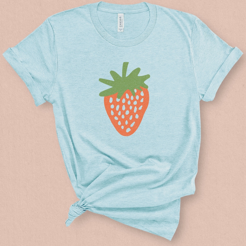 May include: A light blue t-shirt with a stylized orange strawberry graphic. The strawberry has a green stem and leaves.