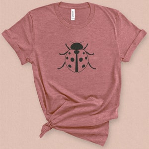 Puede incluir: Una camiseta de color rosa p&aacute;lido jaspeada con una silueta negra de una mariquita en la parte delantera.