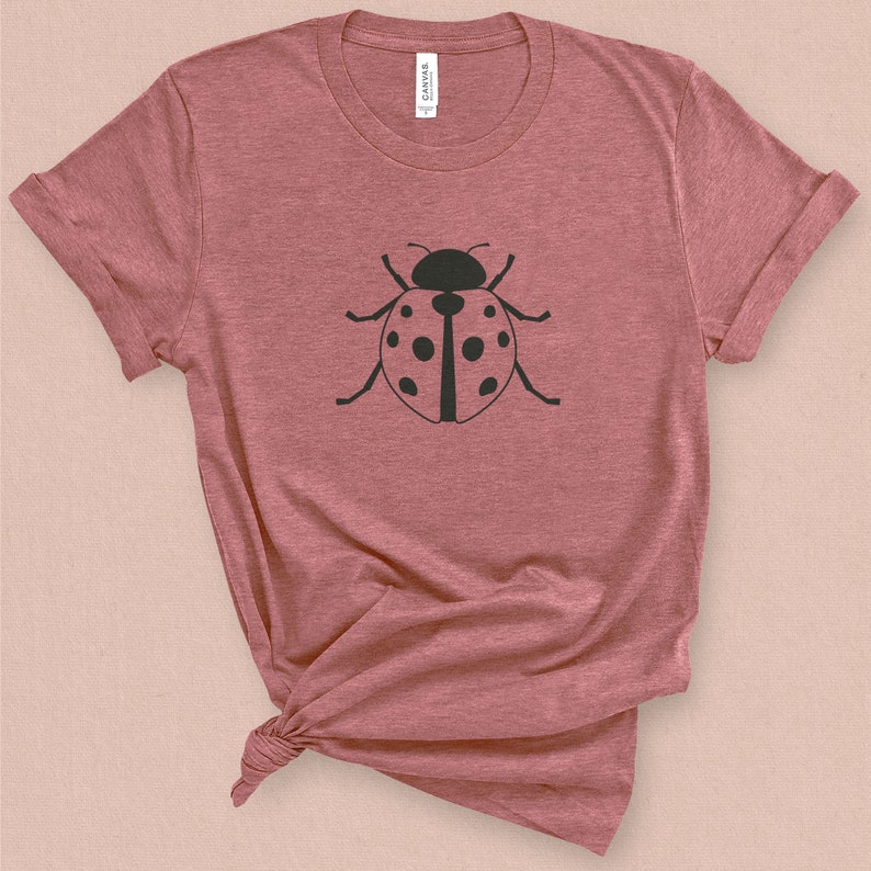 Puede incluir: Una camiseta de color rosa p&aacute;lido jaspeada con una silueta negra de una mariquita en la parte delantera.