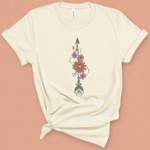 May include: A cream-colored t-shirt with a floral arrow design. The arrow is teal and brown with pink and orange flowers.
