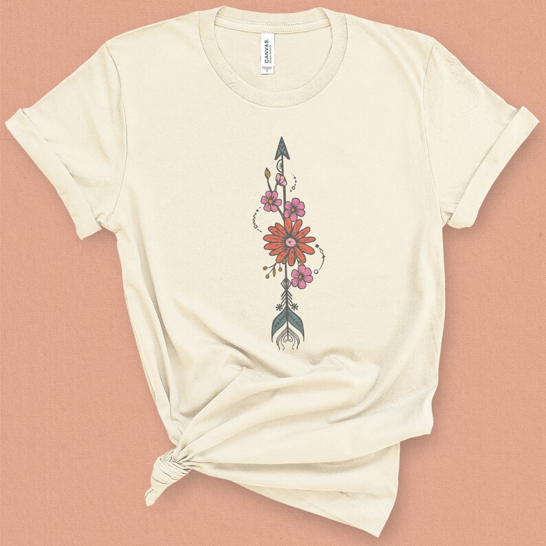 May include: A cream-colored t-shirt with a floral arrow design. The arrow is teal and brown with pink and orange flowers.