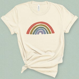 Puede incluir: Una camiseta de color crema con un gráfico de arcoíris impreso en la parte delantera. El arcoíris está formado por rojo, naranja, amarillo, verde, azul y morado.