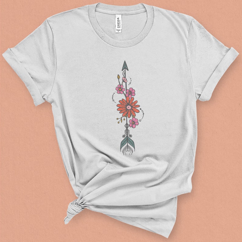 May include: White t-shirt with a floral arrow design. The arrow is green and gray with pink and orange flowers.