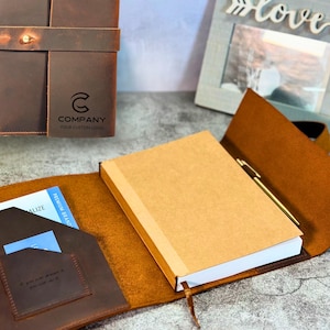 May include: A brown leather journal with a strap closure, a pen, and a notepad. The journal is open, revealing a notepad with a tan cover. The journal has a pocket with a card inside. The text "COMPANY YOUR CUSTOM LOGO" is visible.