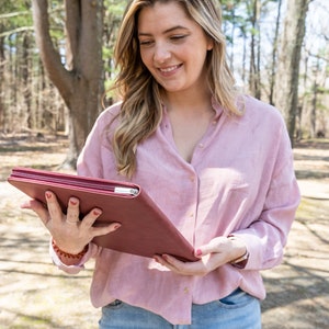 May include: A woman in a pink button-down shirt and blue jeans is holding a burgundy leather planner.