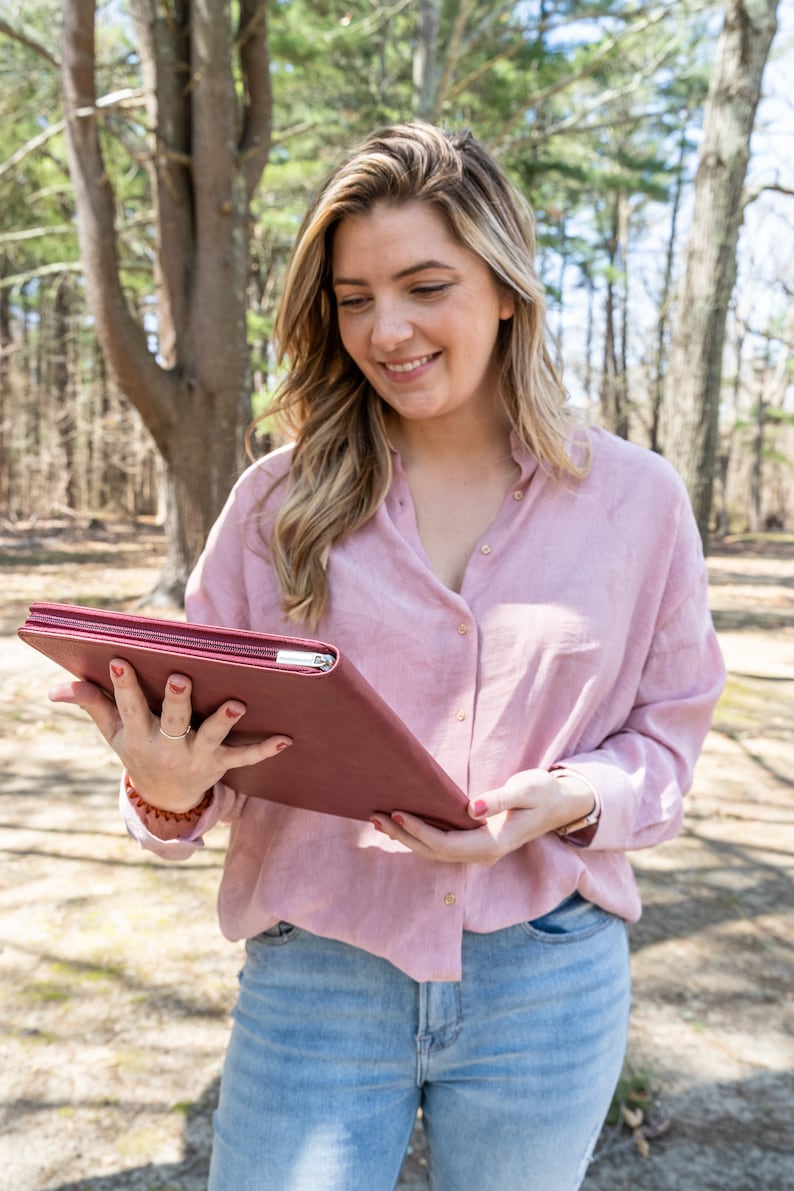 May include: A woman in a pink button-down shirt and blue jeans is holding a burgundy leather planner.