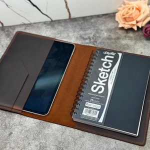 May include: A brown leather notebook cover with a built-in pocket for a tablet or phone. The cover has a black notebook with a spiral binding and a white label that says "Sketch" on it. The notebook is open to a blank page. A gold pen is tucked into the cover.