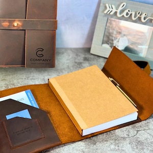 May include: A brown leather journal cover with a wrap-around strap and a metal snap closure. The cover features a debossed logo with the text COMPANY YOUR CUSTOM LOGO. Inside, it holds a tan notebook, a gold pen, and a pocket with a card.