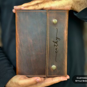May include: A dark brown leather journal cover featuring a vertical flap secured by two silver-toned metal snap buttons. The flap is engraved with the name "cathy" in a cursive script font. The leather has a visible grain and tonal variations.
