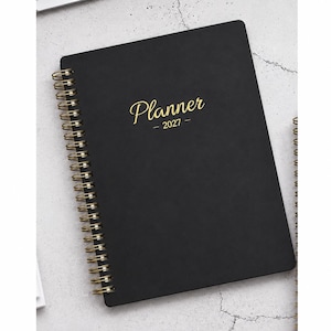 May include: A black spiral-bound planner with gold wire binding. The front cover features the word Planner in a gold script font, with the year 2027 printed in a smaller gold sans-serif font directly underneath the word Planner.