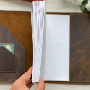 May include: A brown leather journal with a blank white page open. The journal has a pocket on the front cover and a pen holder on the back cover.