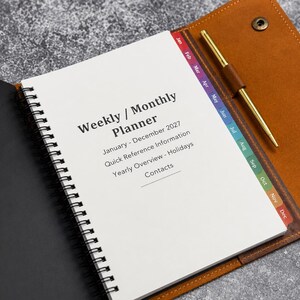 May include: A spiral-bound weekly and monthly planner for January through December 2027, featuring a tan leather cover with a snap closure, a gold pen in a loop, and colorful monthly index tabs. The white pages include sections for holidays and contacts.