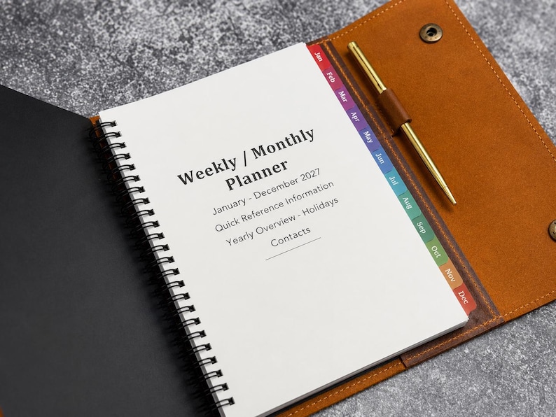 May include: A spiral-bound weekly and monthly planner for January through December 2027, featuring a tan leather cover with a snap closure, a gold pen in a loop, and colorful monthly index tabs. The white pages include sections for holidays and contacts.
