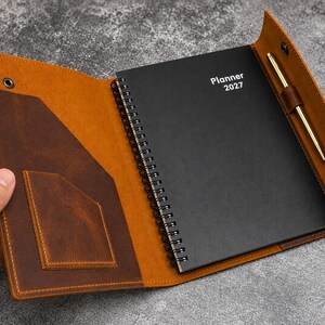 May include: A brown leather planner cover with a snap closure, featuring an interior pocket and a pen loop holding a gold-tone pen. Inside is a black spiral-bound planner with the white text Planner 2027 printed on the front cover.