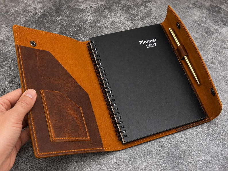 May include: A brown leather planner cover with a snap closure, featuring an interior pocket and a pen loop holding a gold-tone pen. Inside is a black spiral-bound planner with the white text Planner 2027 printed on the front cover.