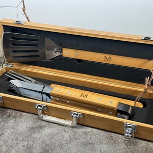May include: A bamboo carrying case with a black interior, containing a spatula and tongs with wooden handles. The handles are engraved with the letter "M".