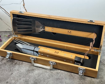 May include: A bamboo carrying case with a black interior, containing a spatula and tongs with wooden handles. The handles are engraved with the letter "M".