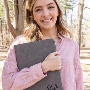 May include: A woman is holding a gray leather portfolio with the initials "R.H." written in black on the front.