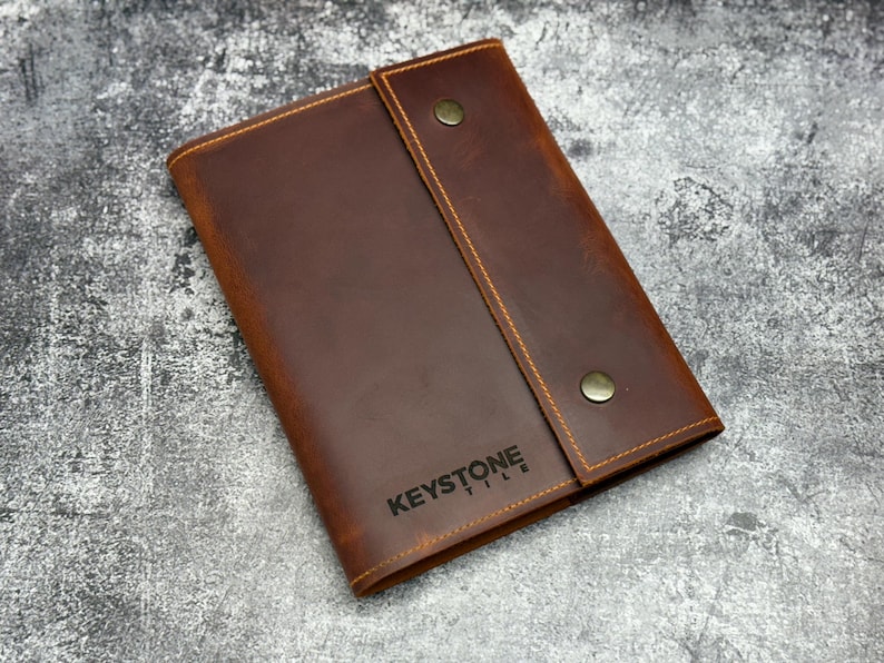 May include: A brown leather notebook cover featuring a vertical flap secured by two brass-colored metal snap buttons. The cover has visible orange stitching along the edges and the text KEYSTONE TILE is embossed in black on the front surface.