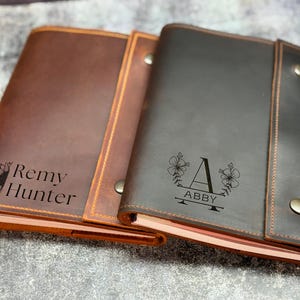 May include: Two rectangular leather journals with snap closures. The left journal is medium brown with a sunflower graphic and the text Remy Hunter. The right journal is dark brown with a floral graphic, the letter A, and the name ABBY.