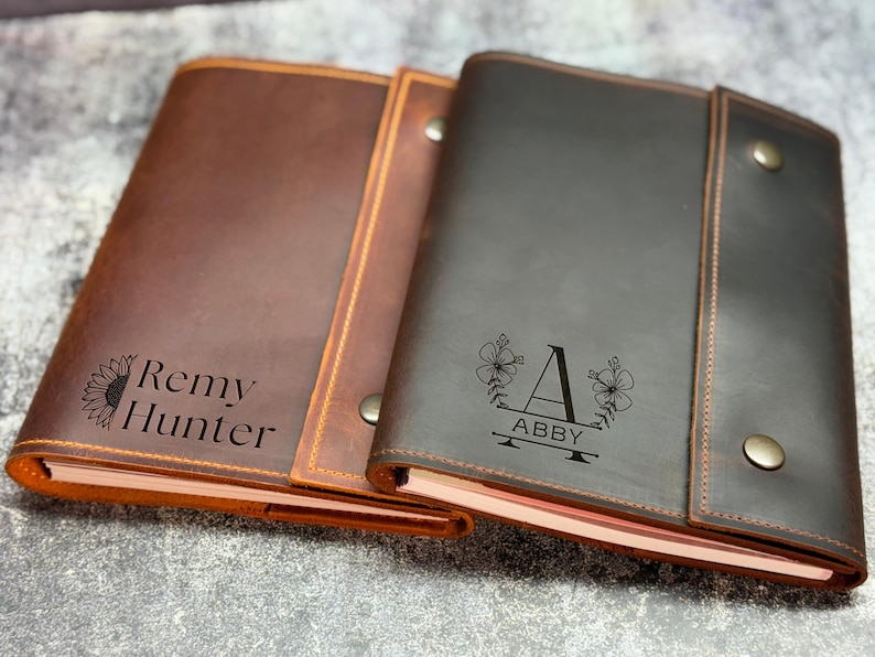 May include: Two rectangular leather journals with snap closures. The left journal is medium brown with a sunflower graphic and the text Remy Hunter. The right journal is dark brown with a floral graphic, the letter A, and the name ABBY.
